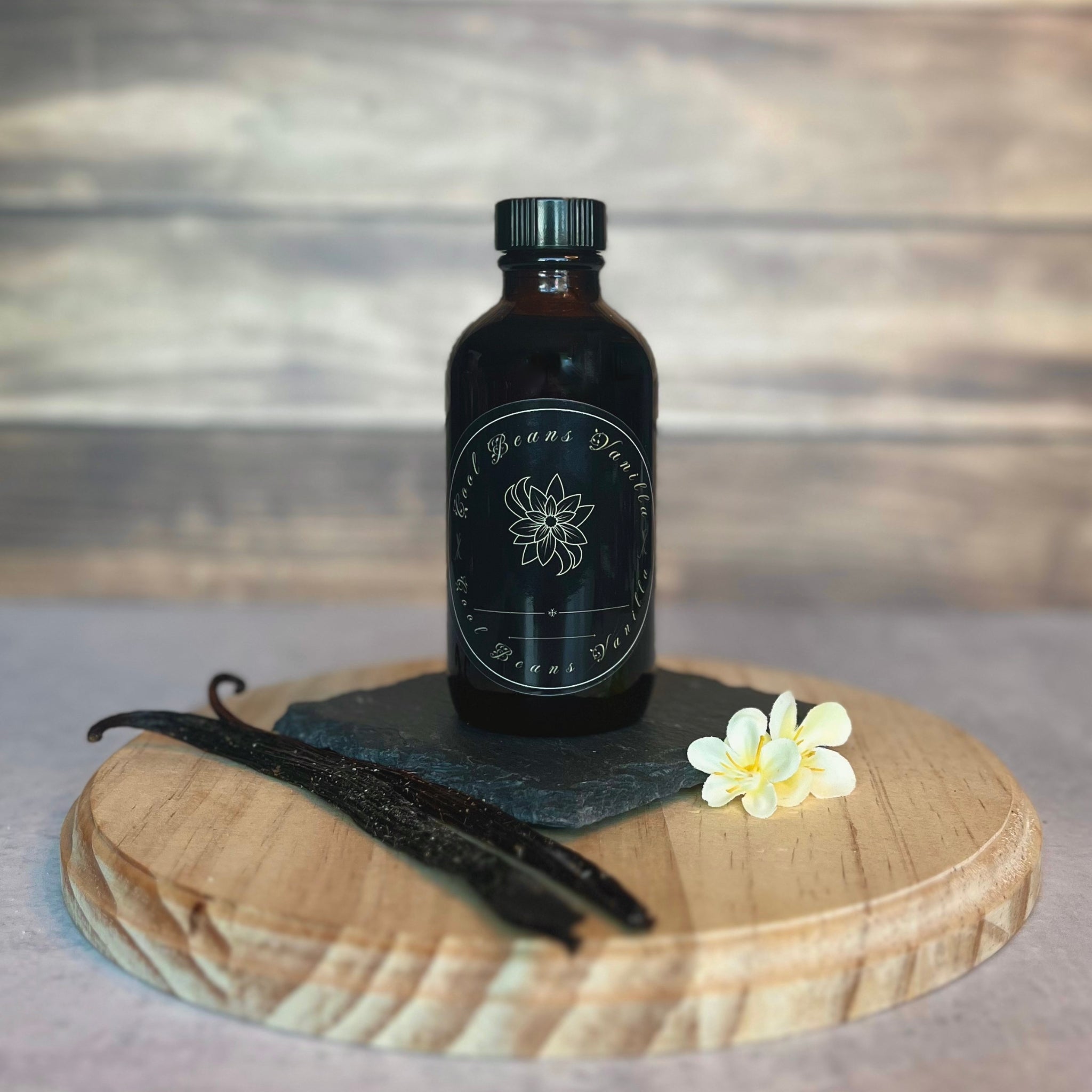 A small, black jar of vanilla extract sits on a dark slate coaster atop a round wooden surface. The jar has a black label with white lettering that reads "Cool Beans Vanilla." Two crystal vanilla pods and a few delicate white flowers are arranged near the base of the jar.