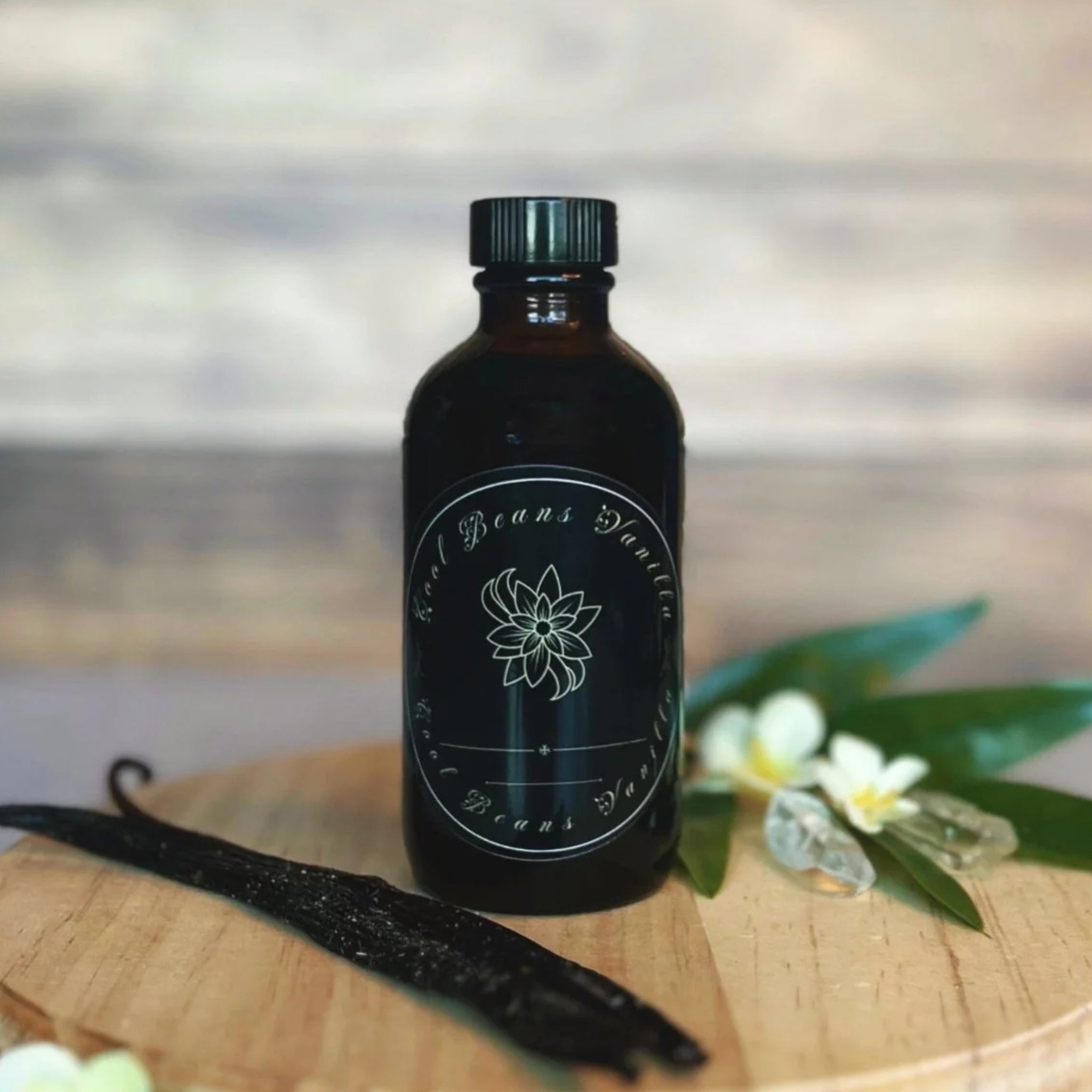 A small, black jar of vanilla extract sits on a round wooden surface. The jar has a black label with white lettering that reads "Cool Beans Vanilla." Two crystal vanilla pods, a few delicate white flowers, and a small quartz crystal are arranged around the base of the jar.