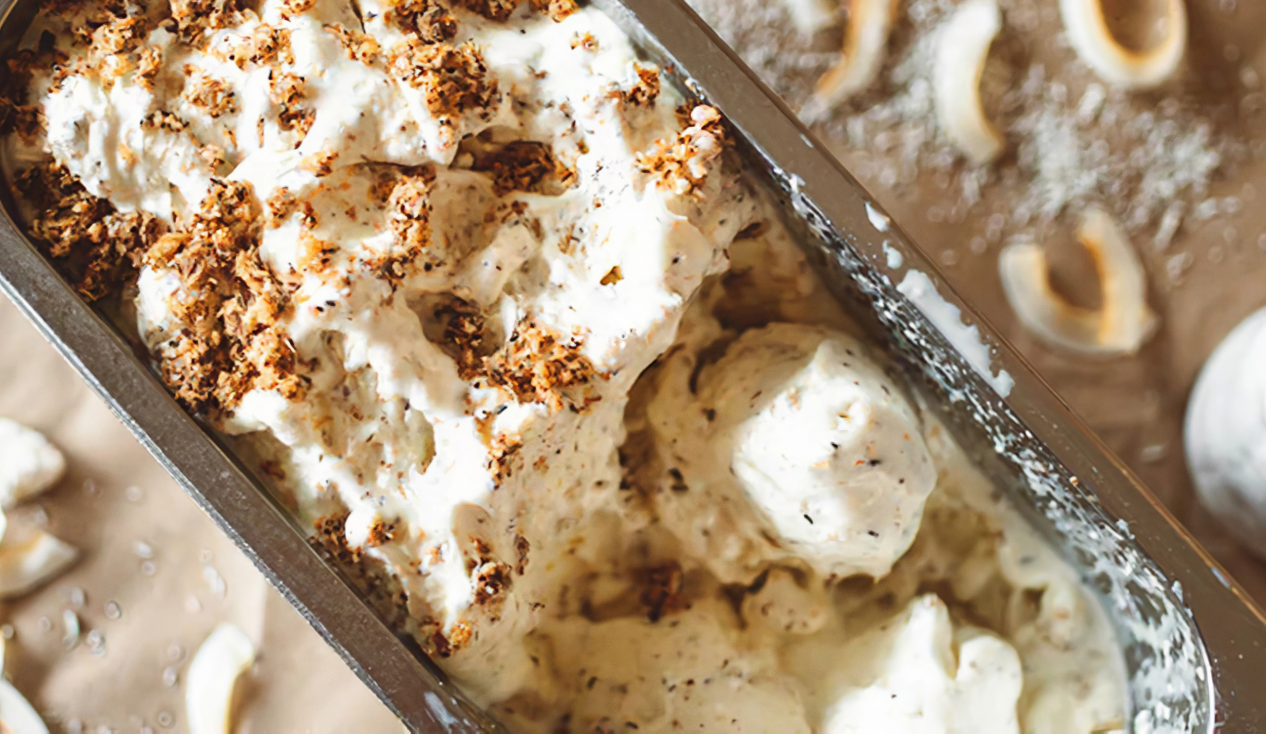 A close-up shot of a metal container filled with creamy white ice cream, generously topped with brown cookie crumbs. Scoops have been taken out of the ice cream, and shavings of coconut are scattered around the container.