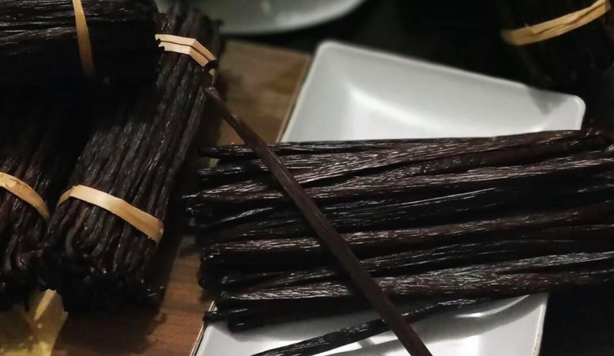 Bundles of dark brown vanilla pods tied with twine, resting on a white rectangular plate and a wooden surface.