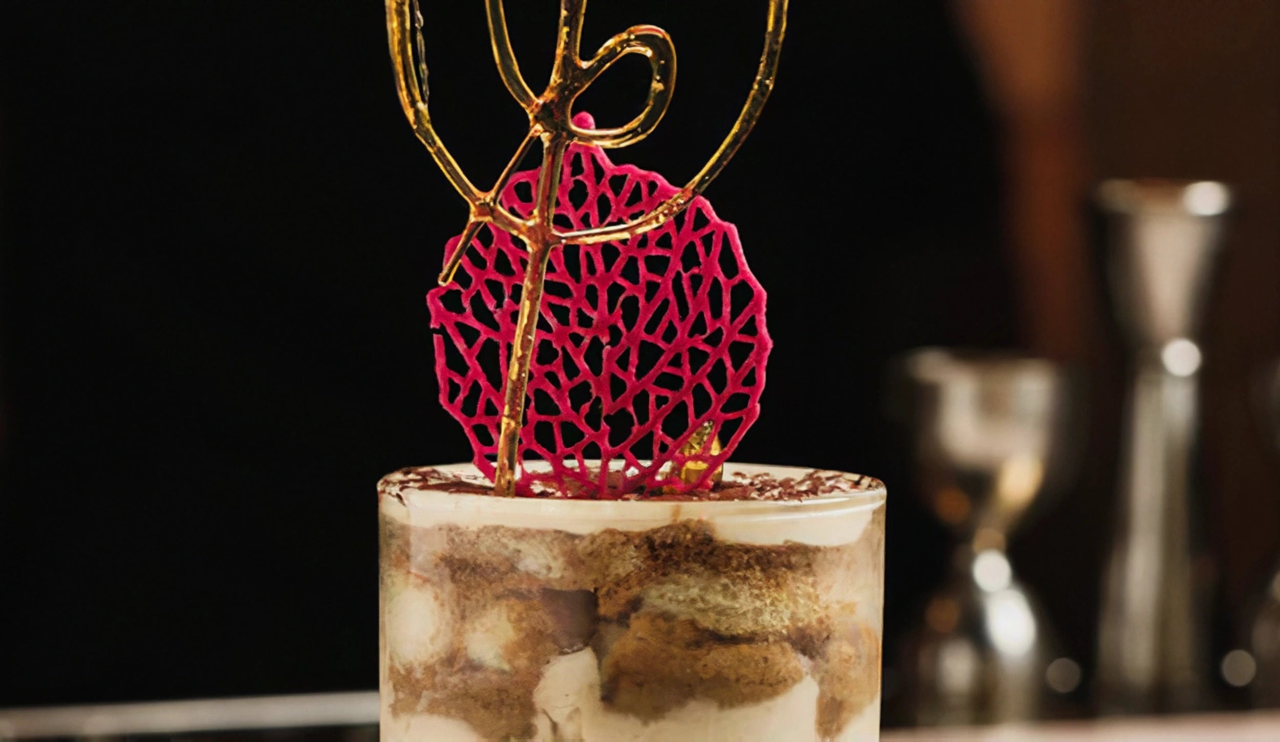 A parfait glass filled with layers of tiramisu dessert. The top layer is dusted with cocoa powder, and the glass is garnished with a pink sugar lace disc and a golden caramel swirl.