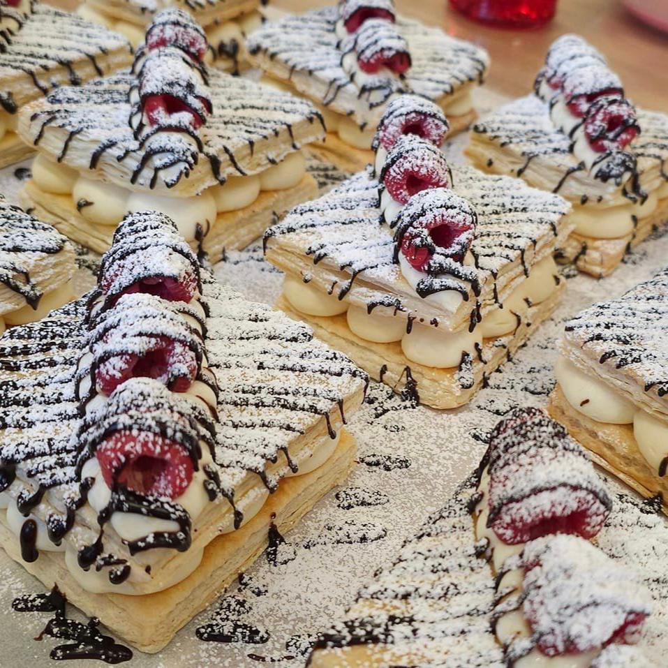 Delicious-looking mille-feuille pastries with golden-brown layers of puff pastry, a creamy filling, and a topping of fresh raspberries, drizzled with chocolate and dusted with powdered sugar.