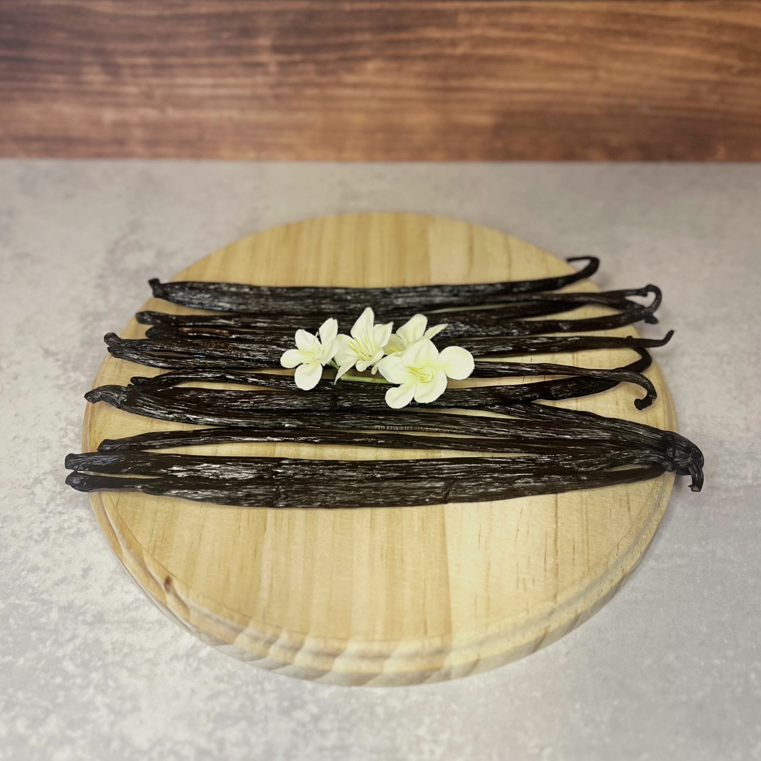 A bundle of dark brown vanilla beans lays on a round wooden surface with a few delicate white flowers resting on top.