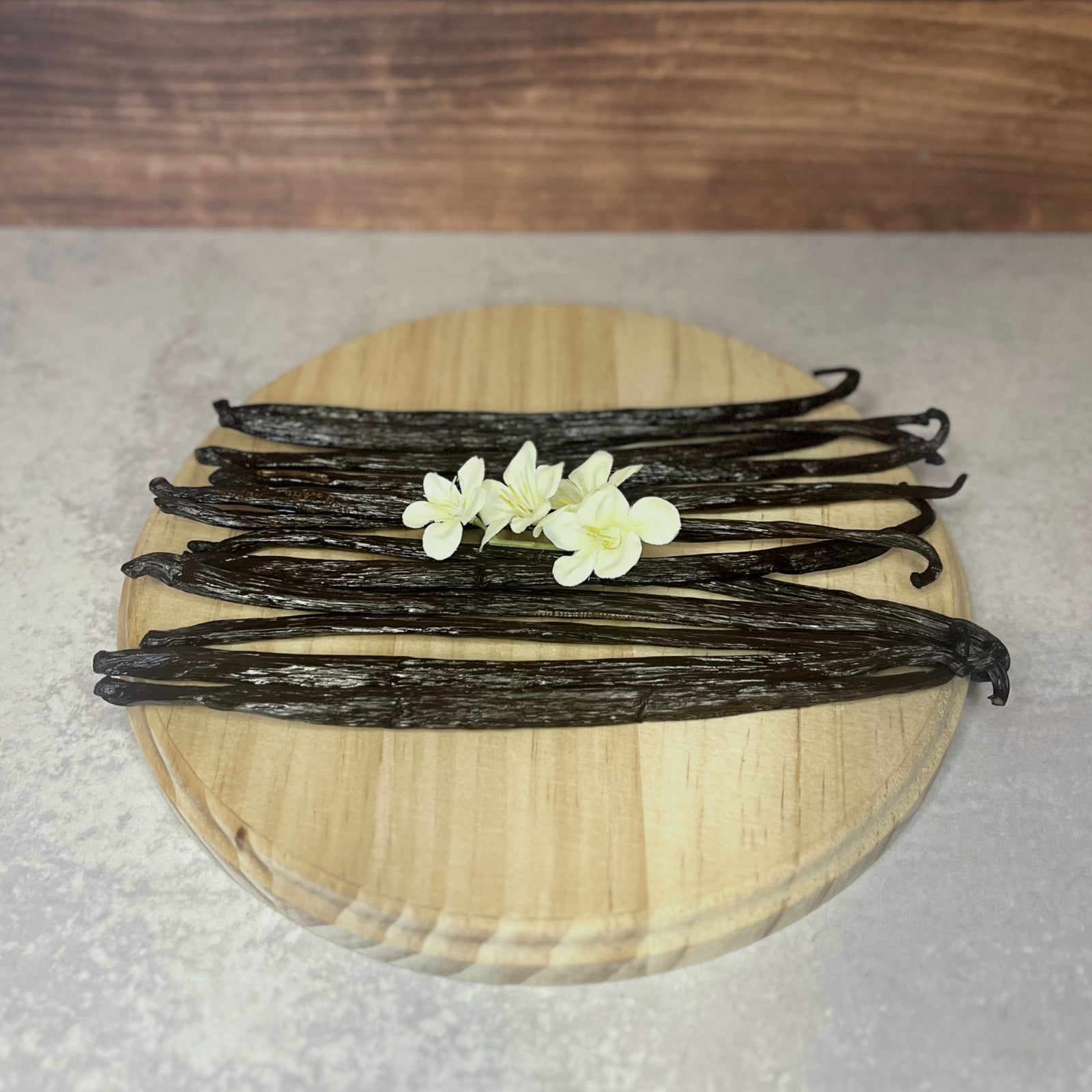 A bundle of dark brown vanilla beans lays on a round wooden surface with a few delicate white flowers resting on top.