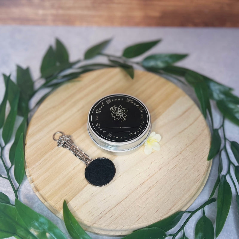 A small, round tin of black vanilla caviar sits on a wooden board, accompanied by a tiny silver spoon with some of the caviar in its bowl. The tin's lid is adorned with a gold-foiled floral design. A few green leaves and a small yellow flower are scattered around the board.