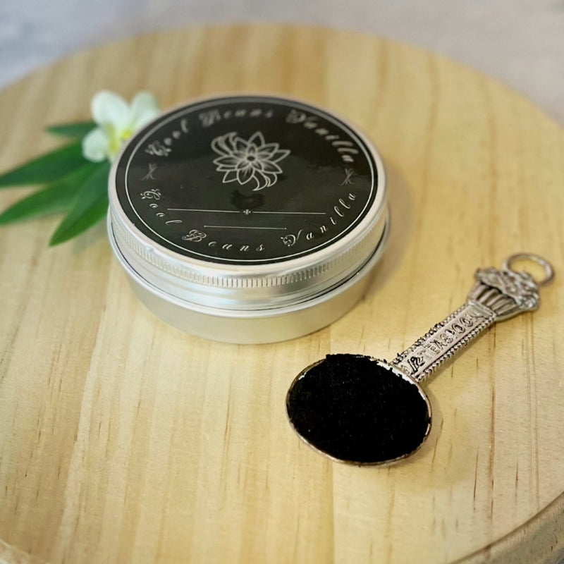 A small tin of "Vanilla Caviar Seeds" is placed on a wooden board. A small spoon next to it holds a portion of the seeds, showing their fine, dark-colored texture.