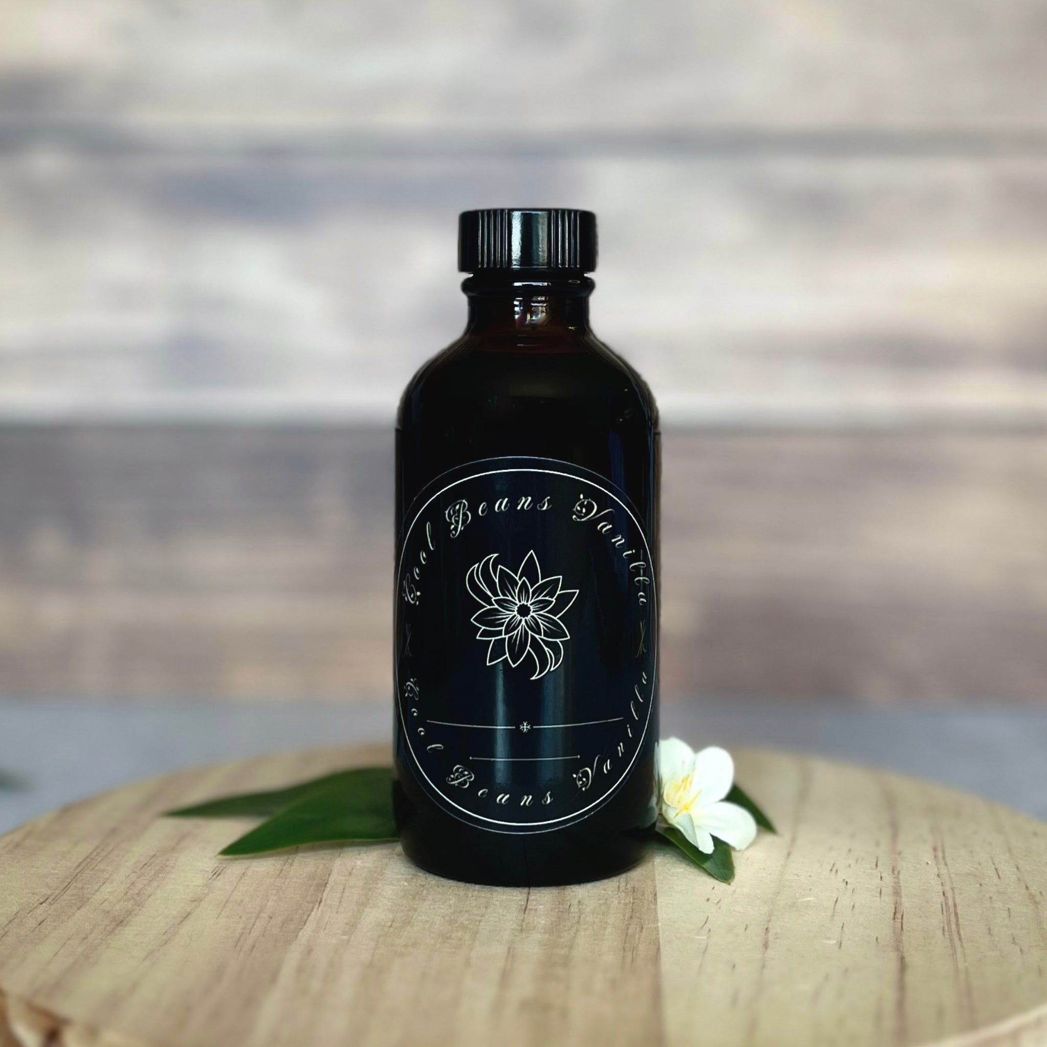 Close-up of a dark glass bottle of vanilla extract with a black screw cap and a white label showing a stylized flower and the text "Cool Beans Vanilla." The bottle sits on a round, light-colored wooden board against a blurred wooden backdrop. A white flower and green leaves are positioned near the bottle.