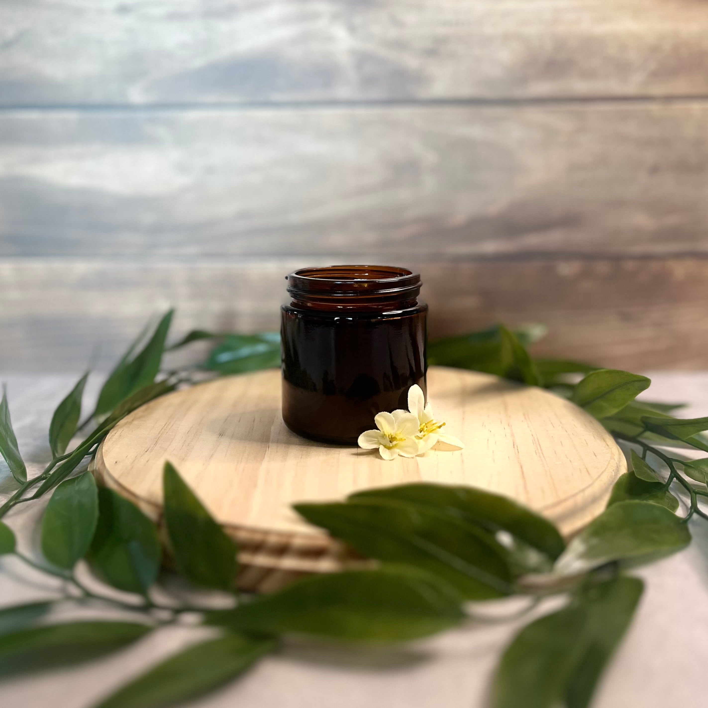 A small, amber glass jar sits on a round wooden surface.  A few delicate white flowers and green leaves are arranged around the base of the jar. The jar is filled with bourbon vanilla powder.