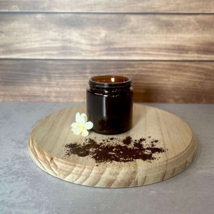 A small, amber glass jar sits on a round wooden surface.  A small pile of dark brown vanilla powder is spilled in front of the jar, and a few delicate white flowers are arranged near the base of the jar.