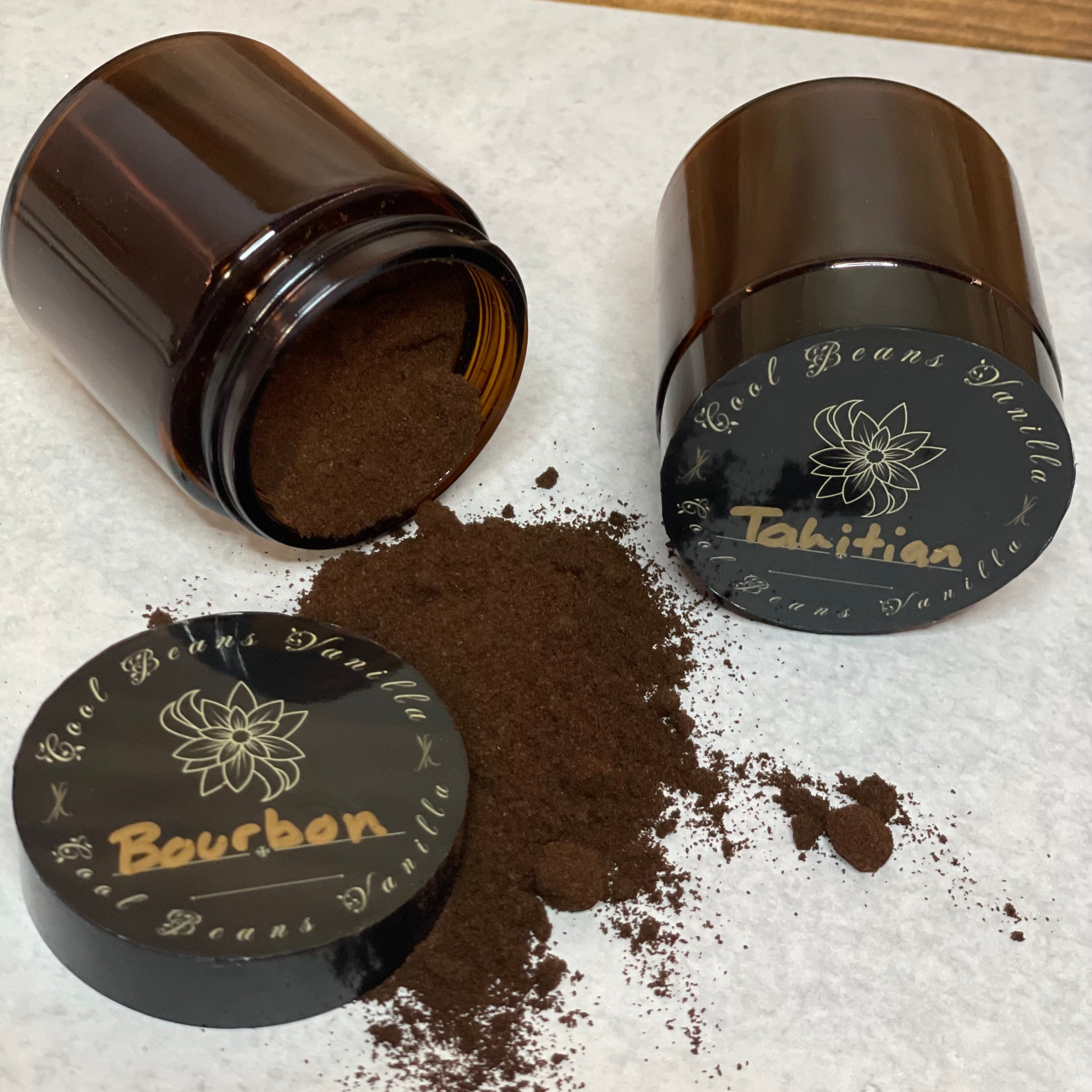 Two amber glass jars lay on their sides with their lids off, spilling dark brown vanilla powder onto a white surface. One lid is labeled "Bourbon" and the other "Tahitian," indicating the different types of vanilla powder.