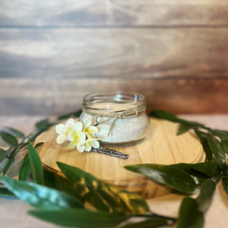 A small glass jar filled with sugar scrub sits on a wooden surface. The jar is decorated with a piece of twine and a small artificial flower. A vanilla bean lies next to the jar.