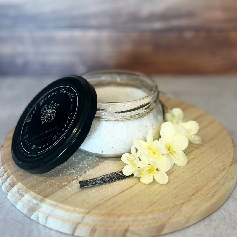 A small, open glass jar of vanilla sugar scrub sits on a wooden board, with the lid next to it.  A vanilla bean and a few small yellow flowers are arranged near the jar.