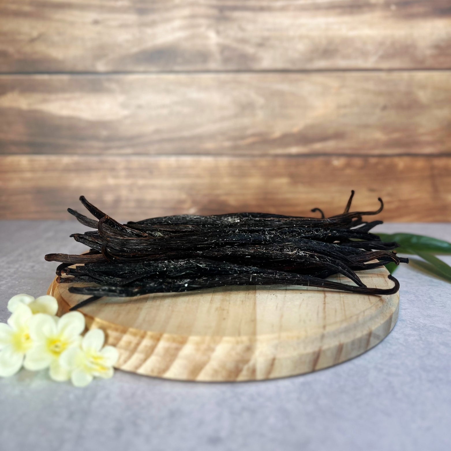 A bundle of plump, dark brown vanilla beans with crystals throughout them, sits on a round wooden surface. A few delicate white flowers are arranged near the base of the wooden surface.