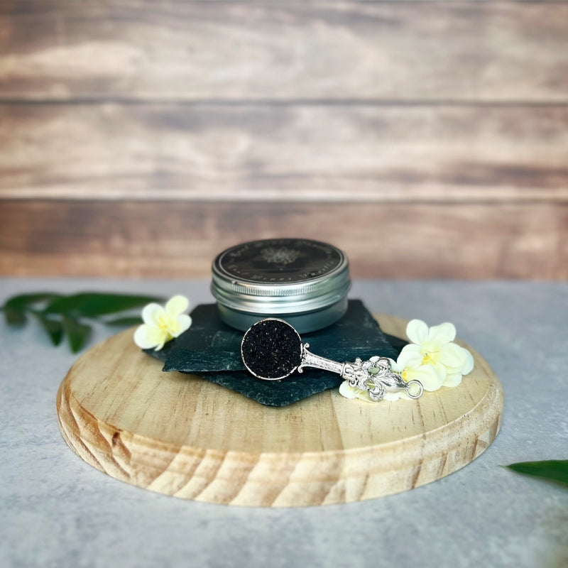 A small, silver tin of vanilla caviar seeds sits on a dark stone slab atop a round wooden surface.  A decorative silver spoon filled with the tiny black vanilla seeds rests on top of the tin.  A few delicate white flowers are arranged near the tin.