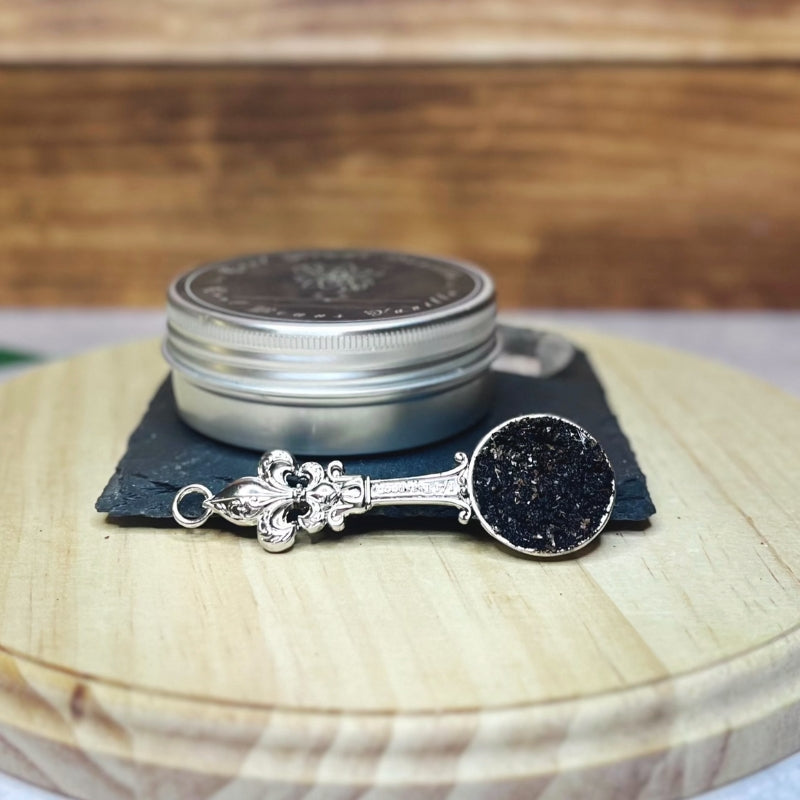 A small, silver tin of vanilla caviar seeds sits on a dark slate coaster on a round wooden surface. In the foreground, an ornate silver spoon with a fleur-de-lis on the handle is filled with the tiny black vanilla seeds with small crystals.