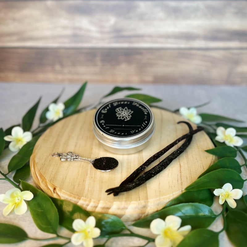 A small, silver tin of vanilla caviar seeds sits on a round wooden surface. Beside it, two crystal vanilla pods and a small, ornate silver spoon filled with the tiny black vanilla seeds are arranged.  Delicate white flowers and green leaves encircle the wooden surface.