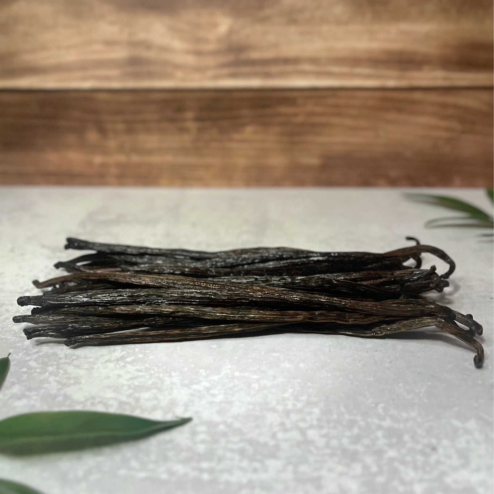 A bundle of Grade B vanilla beans lays on a gray surface. The beans are a dark brown color and are slightly drier than Grade A. Green leaves peek in from the sides of the image.