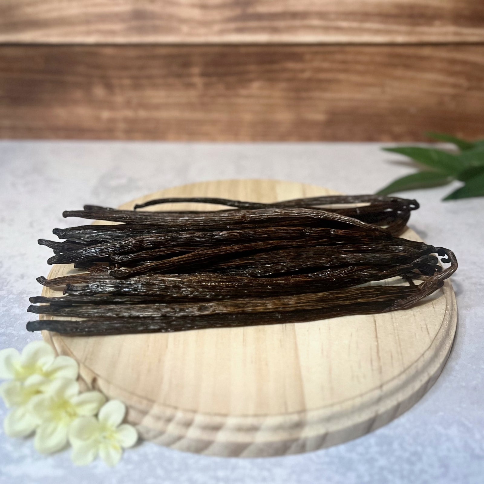 A bundle of Grade B vanilla beans lays on a round wooden surface. The beans are a dark brown color and have a slightly dry appearance.  A few delicate white flowers are arranged near the base of the wooden surface.