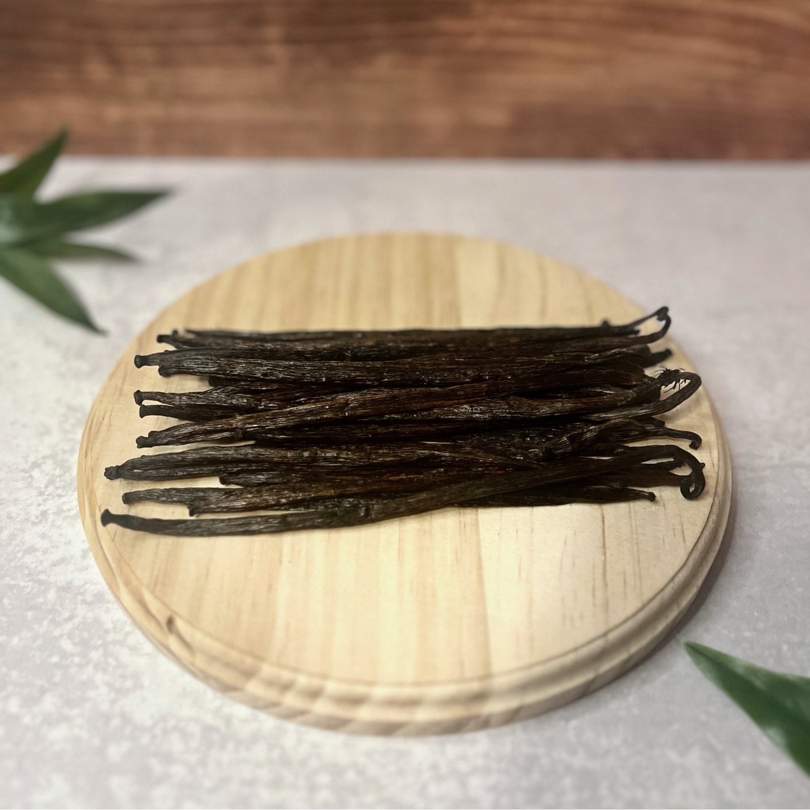 A bundle of Grade B vanilla beans lays on a round wooden surface. The beans are a dark brown color and have a slightly wrinkled and uneven appearance, which is typical of this grade.