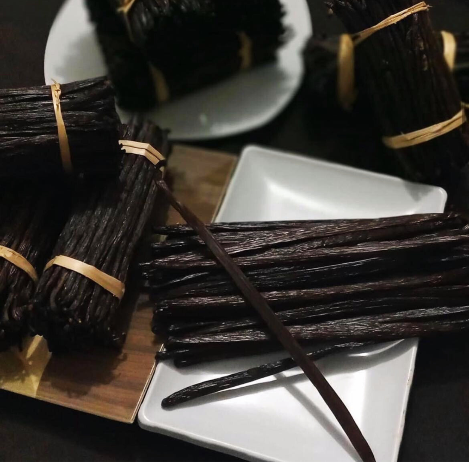 Several bundles of vanilla beans are arranged on a table. The vanilla beans are tied together with twine and vary in size and color. Some bundles are neatly stacked on a wooden surface, while others are scattered loosely on a white plate. A close-up reveals the intricate details of individual vanilla beans, showcasing their rich brown hue and wrinkled texture.