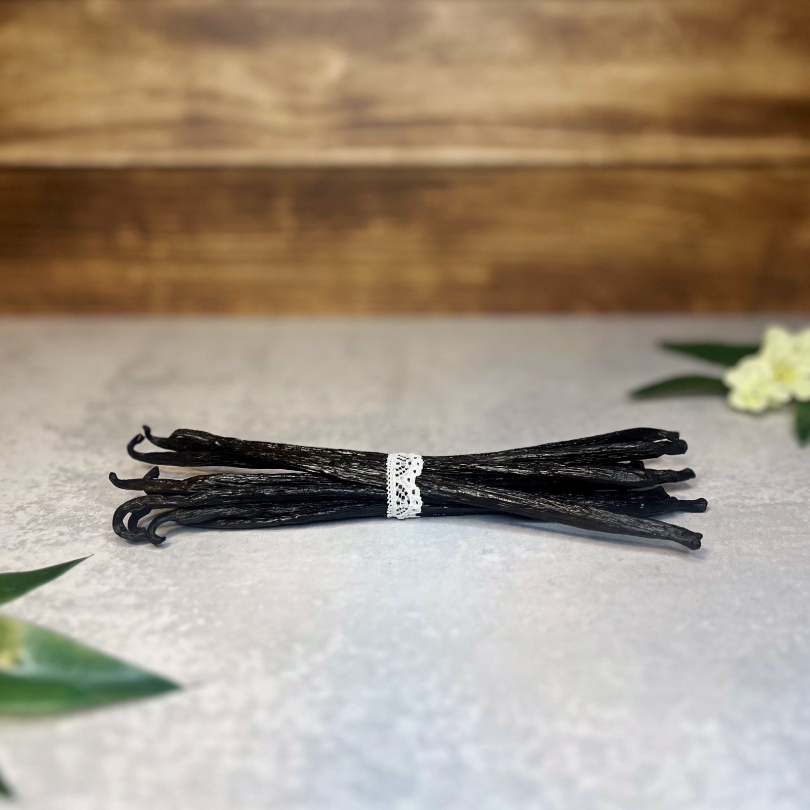 A bundle of dark brown vanilla beans tied with a white ribbon lays on a gray surface.  A few delicate white flowers and green leaves are arranged near the bundle.