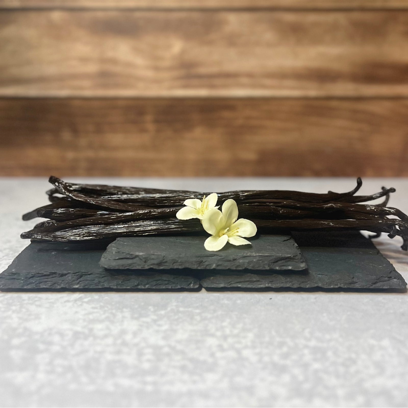 A bundle of dark brown vanilla beans sits atop three stacked dark gray slate coasters. A few delicate white flowers are arranged on the top coaster with the vanilla beans.