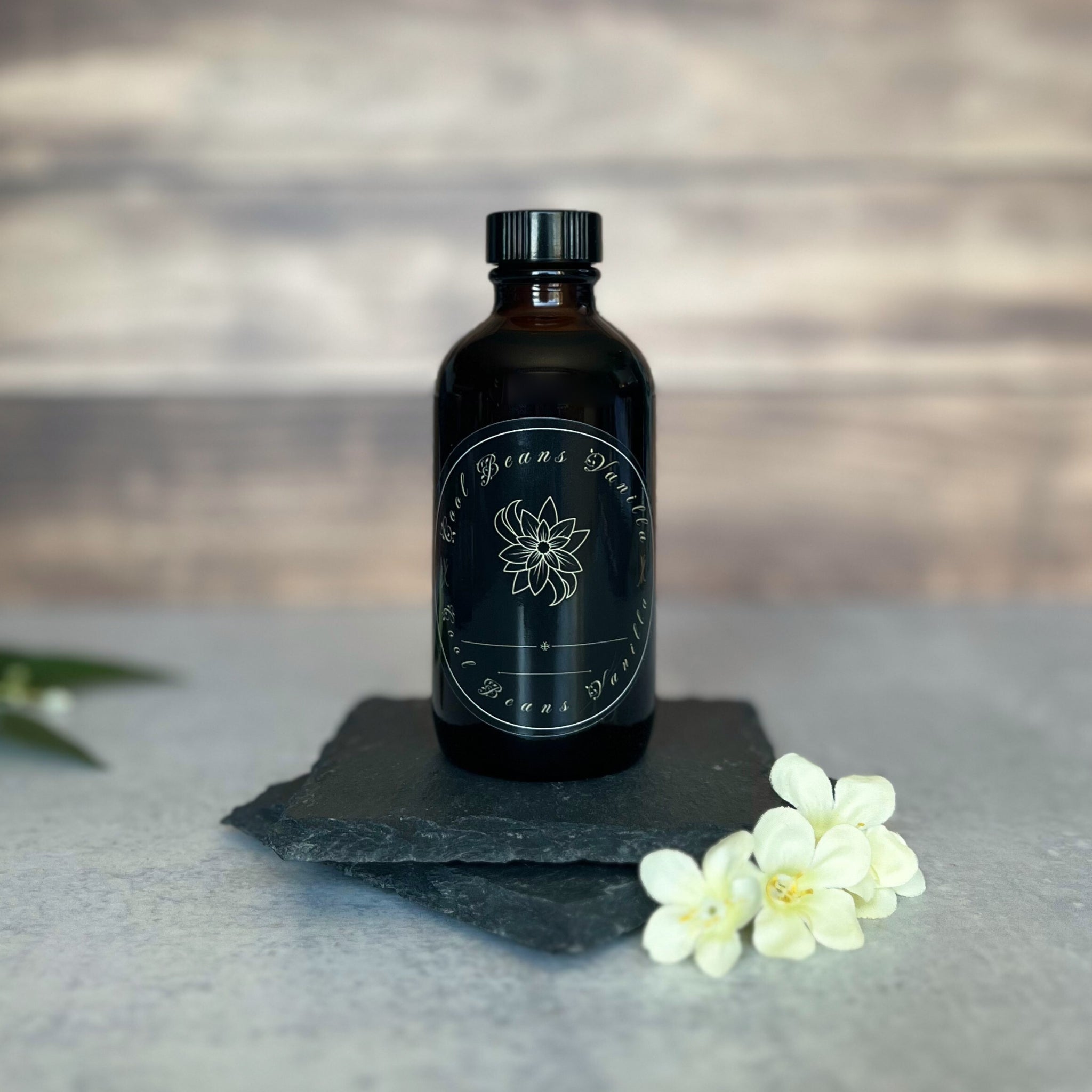 A dark amber glass bottle of "Cool Beans Vanilla" extract with a black label featuring a white floral design and the brand name. The bottle rests on two stacked pieces of dark slate, with a blurred rustic wood background. White flowers are visible in the foreground.