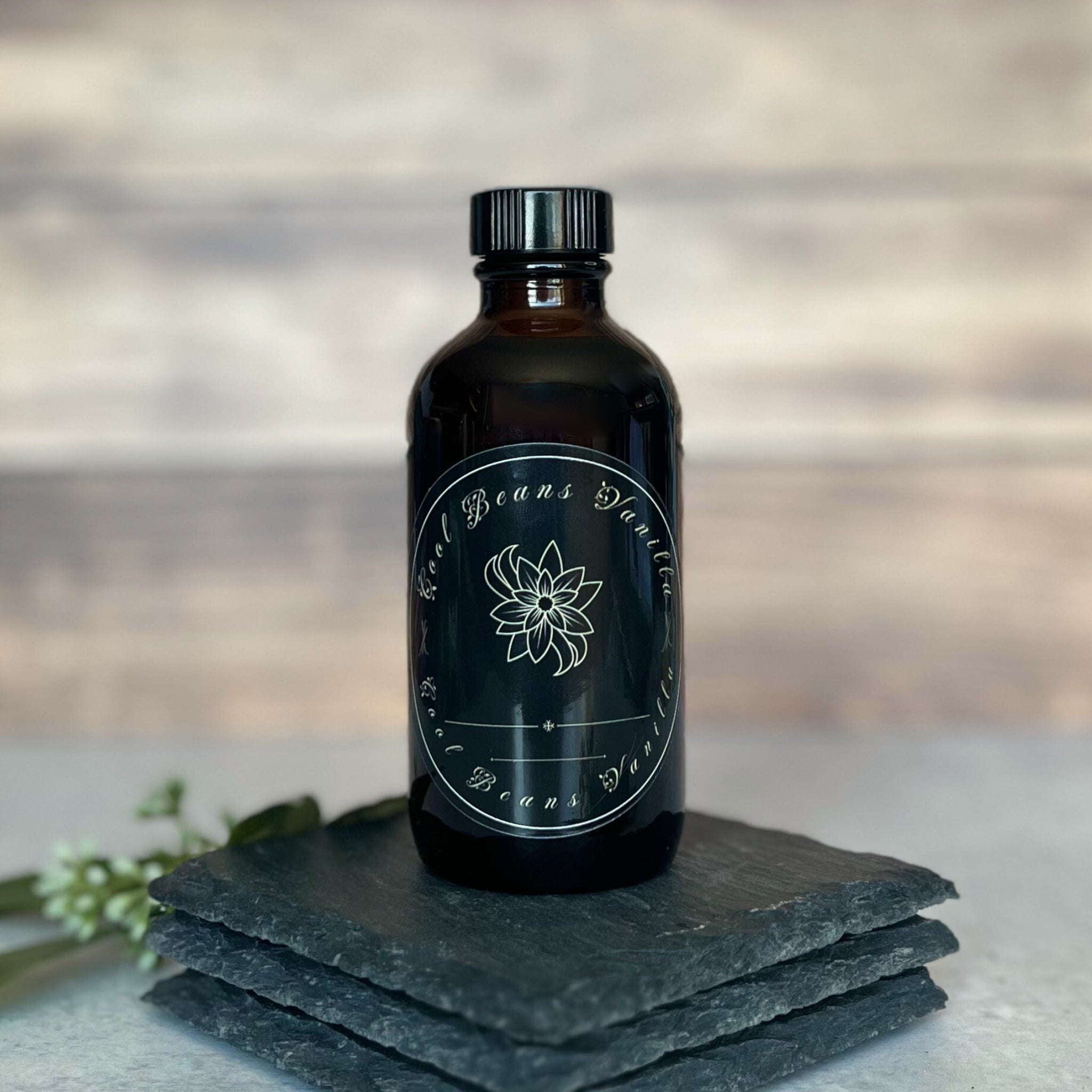 A dark amber glass bottle of "Cool Beans Vanilla" extract with a black label featuring a white floral design and the brand name. The bottle rests on a stack of three dark, rough-textured slate pieces. A blurred rustic wood background is visible, and partially obscured white flowers are in the lower left corner.