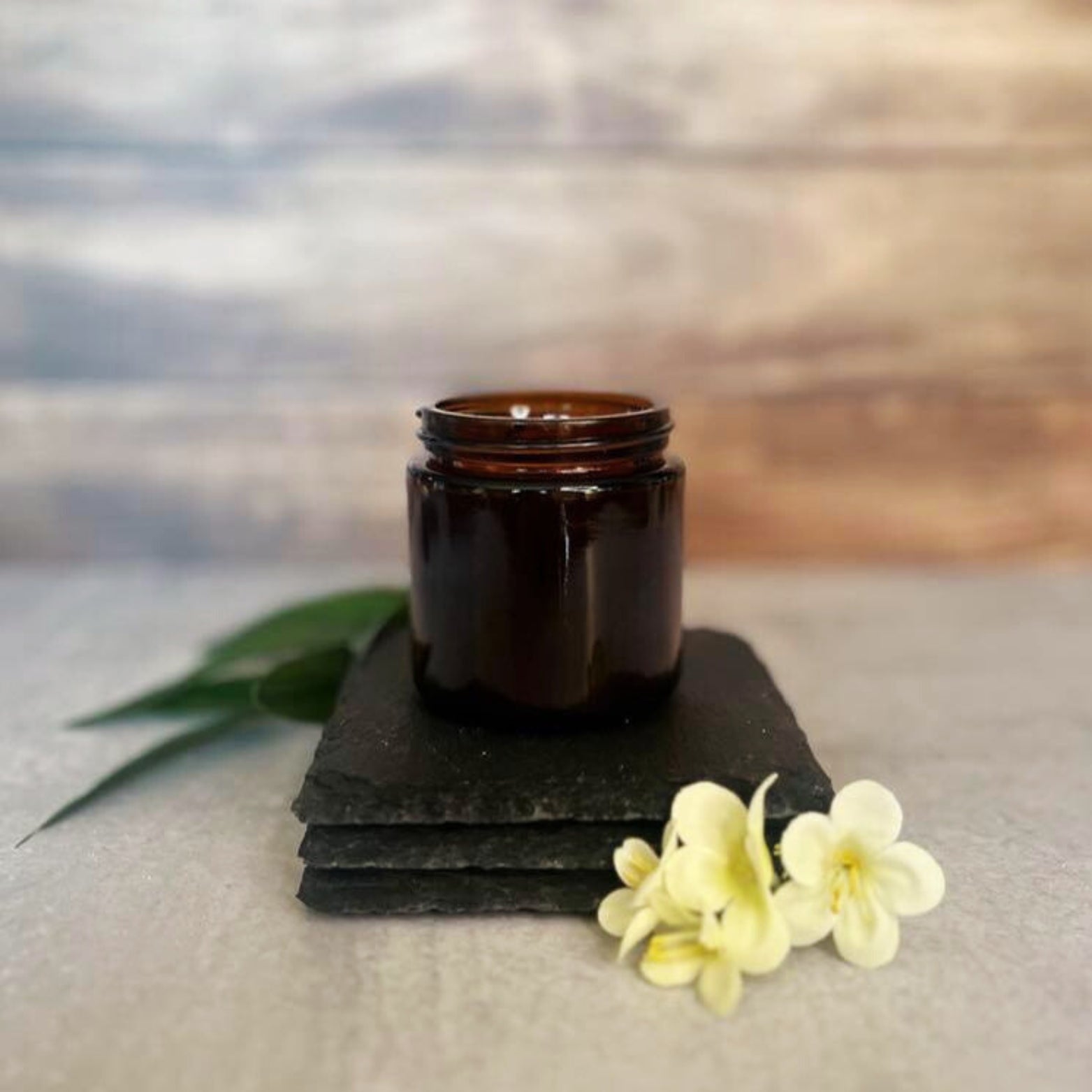 A small, amber glass jar sits on three stacked dark gray slate coasters. A few delicate white flowers are arranged near the base of the coasters. The jar is filled with vanilla powder.