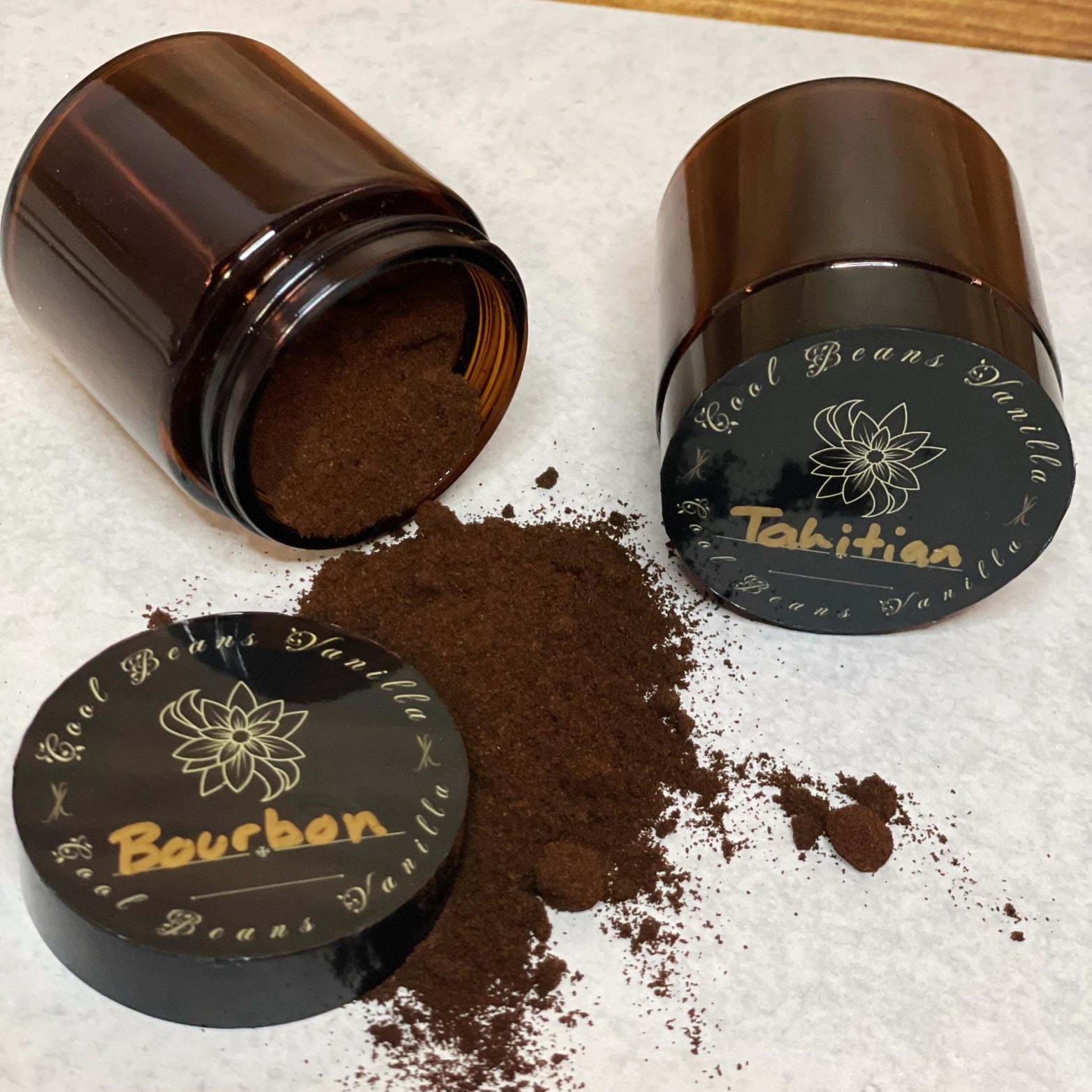 Two amber glass jars lay on their sides with their lids off, spilling dark brown vanilla powder onto a grey surface. One lid is labeled "Bourbon" and the other "Tahitian," indicating the different types of vanilla powder.