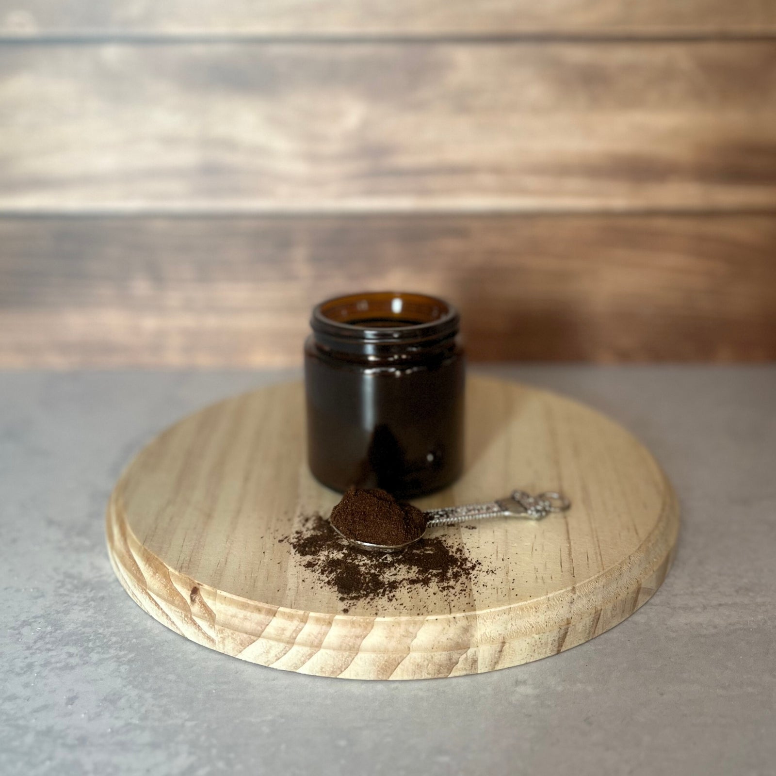 A small, amber glass jar sits on a round wooden surface. In front of the jar, a small pile of dark brown vanilla powder rests on an ornate silver spoon.