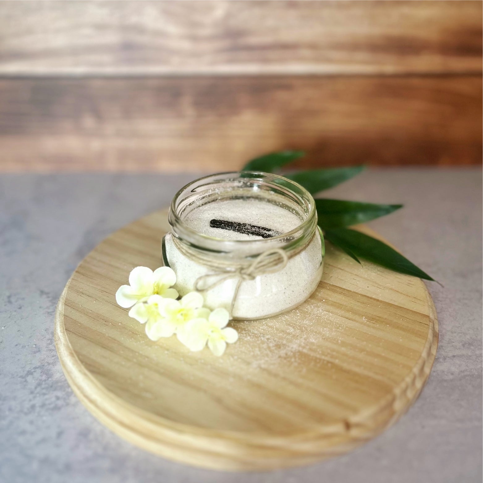 A small glass jar of sugar sits open on a wooden board. A vanilla bean rests inside, and the jar is tied with twine.  A few small white flowers are arranged near the jar.