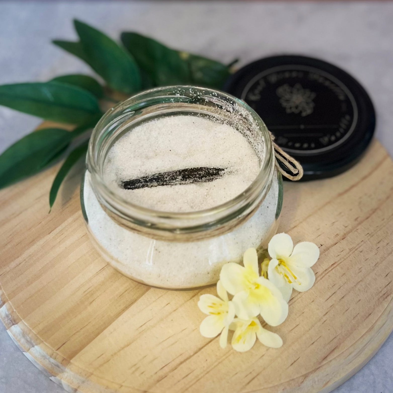 An open jar of vanilla sugar sits on a wooden board, with a vanilla bean visible inside. The lid is placed off to the side, and a few small yellow flowers are arranged near the jar.