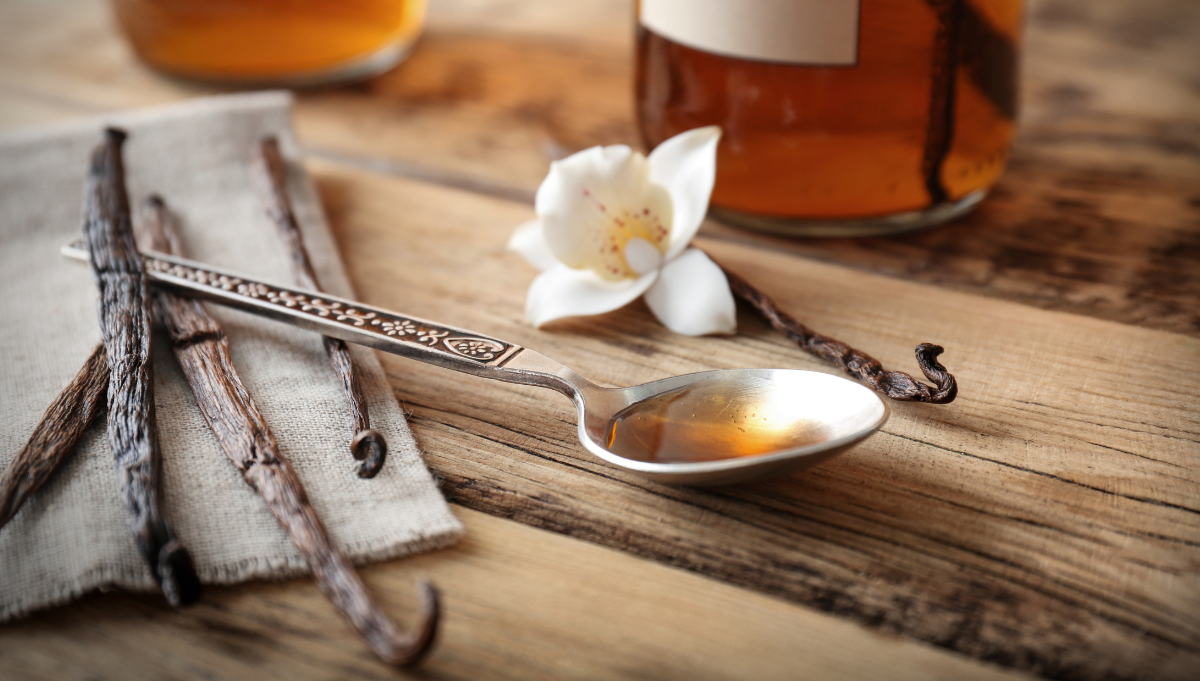 A rustic scene with a spoon holding vanilla extract, surrounded by vanilla beans, a vanilla flower, and a bottle of extract on a wooden table.