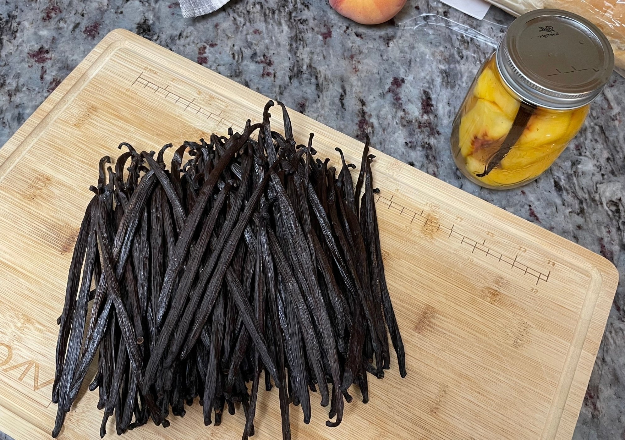 A pile of dark brown vanilla beans sits on a wooden cutting board next to a jar of sliced peaches and vanilla pods.