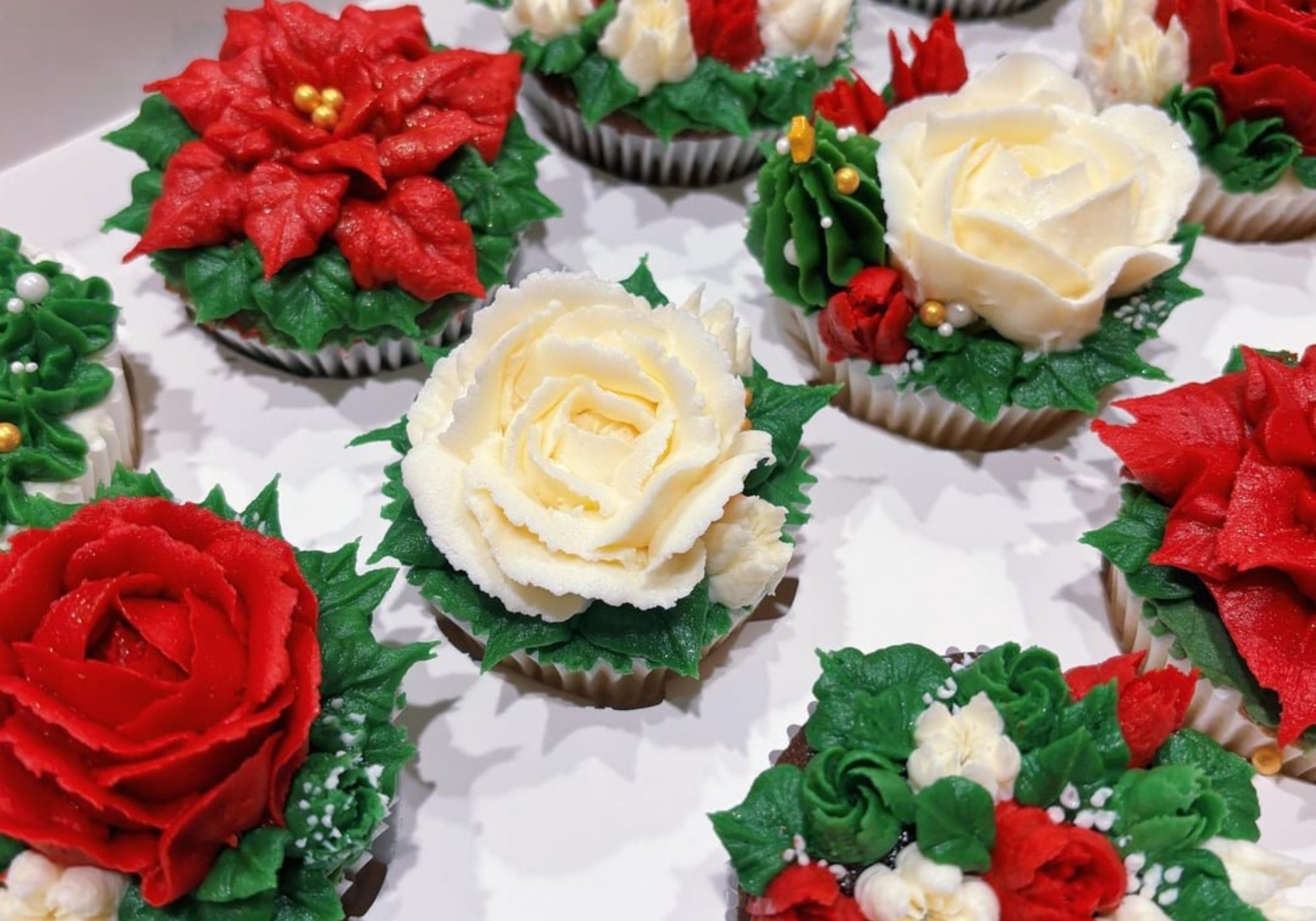 A box filled with cupcakes decorated with elaborate frosting flowers in red, white, and green, resembling poinsettias, roses, and holly.