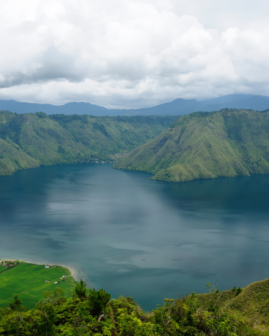 A serene, wide-angle view of a large lake nestled amidst rolling green hills, under a soft, cloudy sky.