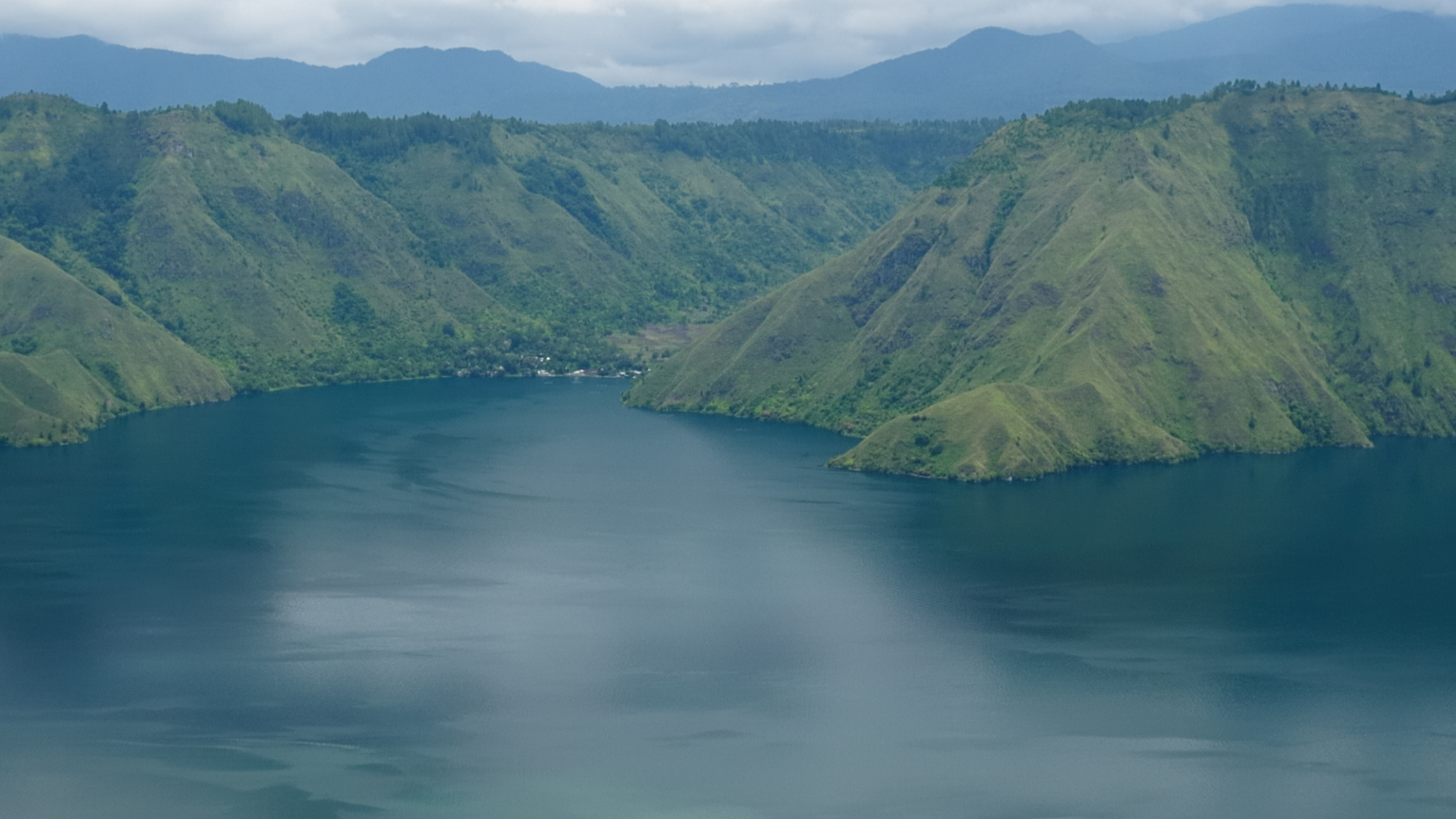 A wide aerial shot of Lake Toba in Sumatra, Indonesia. Steep, green volcanic hills surround the calm, blue water.