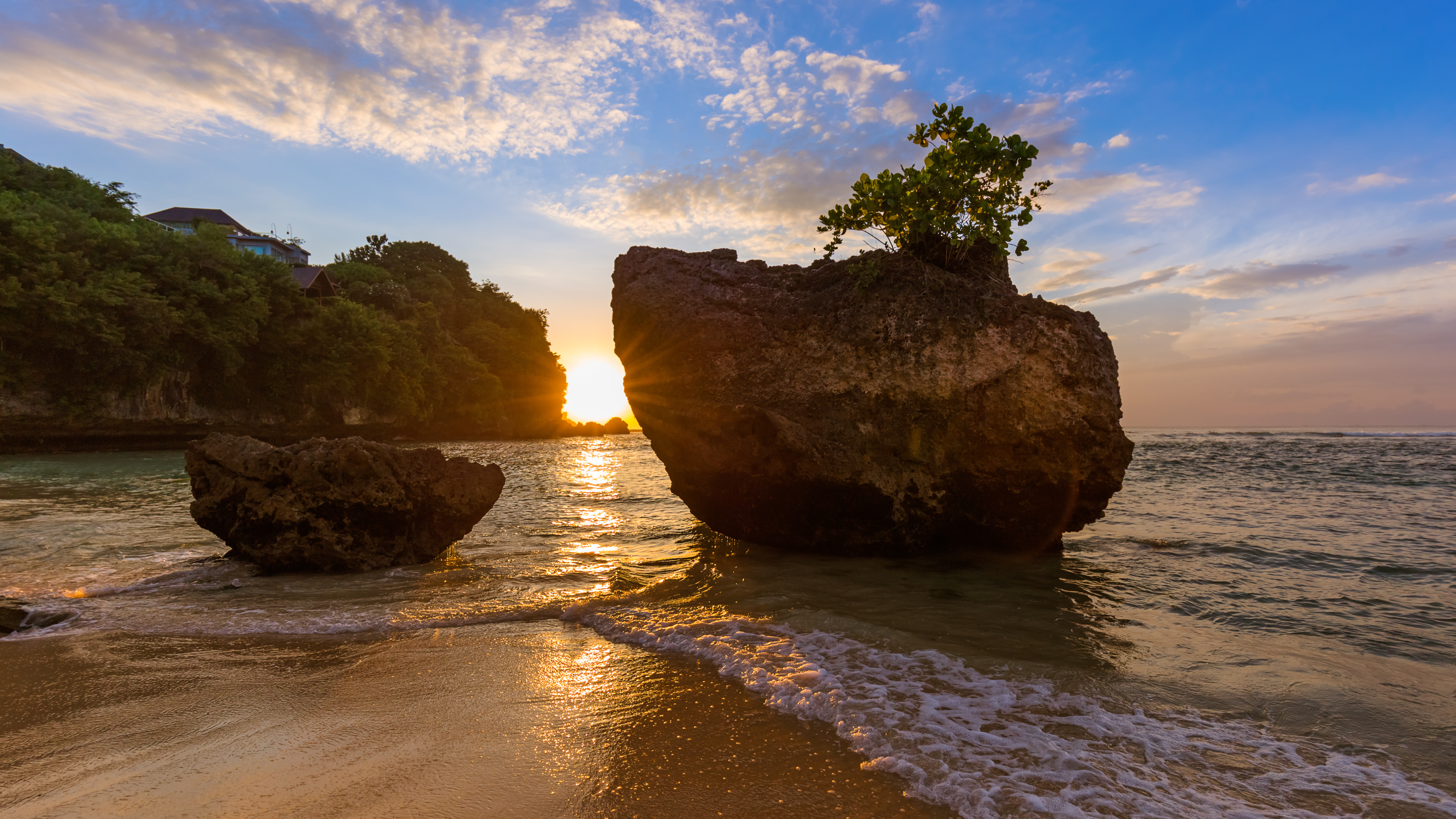 Golden sunlight streams through a gap between two large rocks on a beach at sunset, casting long shadows on the sand and water. Lush green vegetation tops the cliffs in the background.