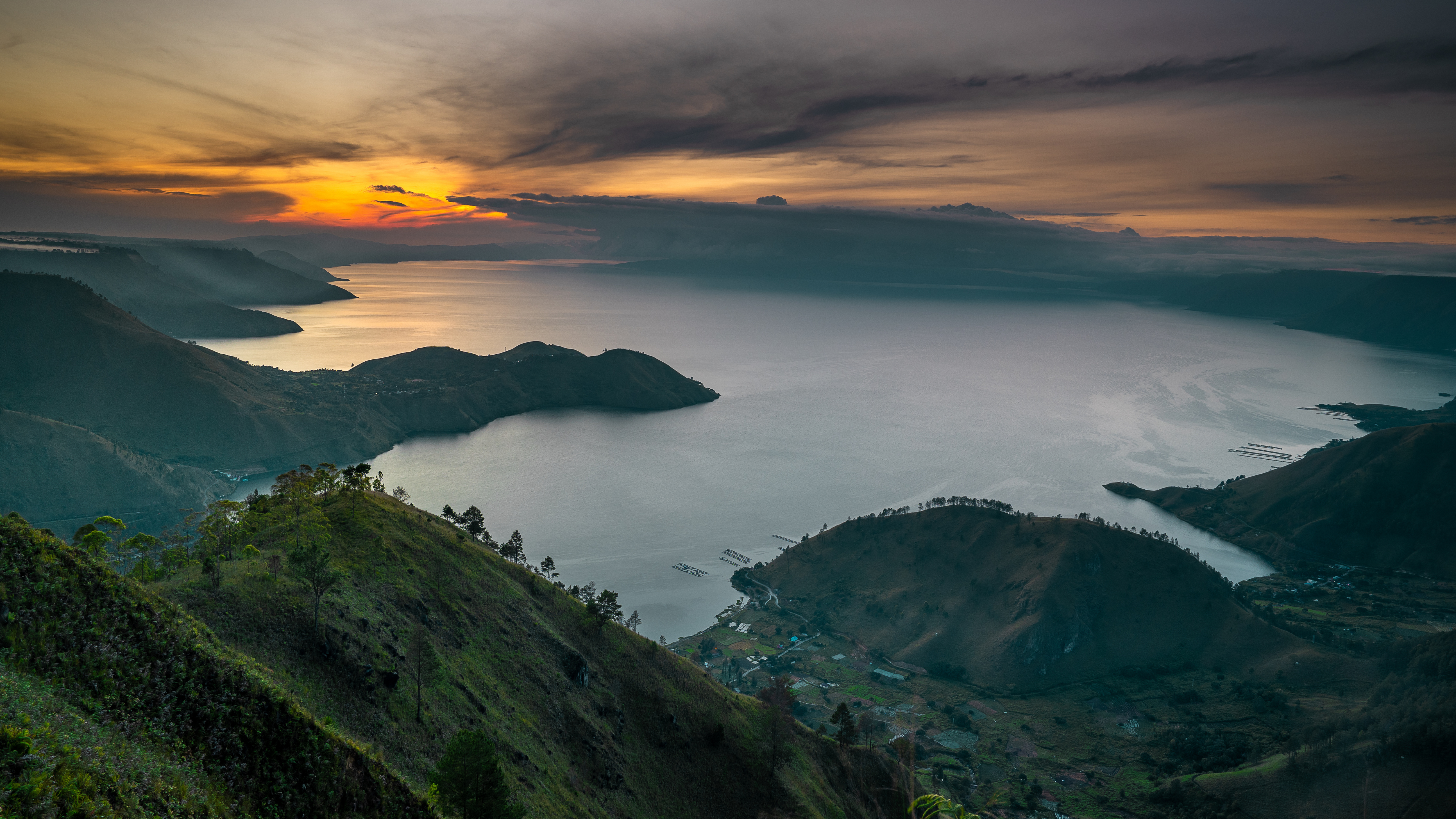 A panoramic view of a vast lake surrounded by rolling hills, with the sun setting dramatically in the background.