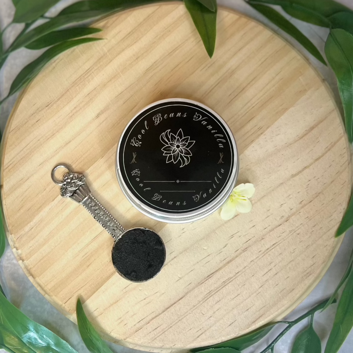 A flat lay product shot featuring a tin of "Cool Beans Vanilla" powder placed on a circular wooden board. An antique-style spoon holds a small amount of the dark brown vanilla powder, showcasing its fine texture.  Green leaves frame the board, adding a natural touch.