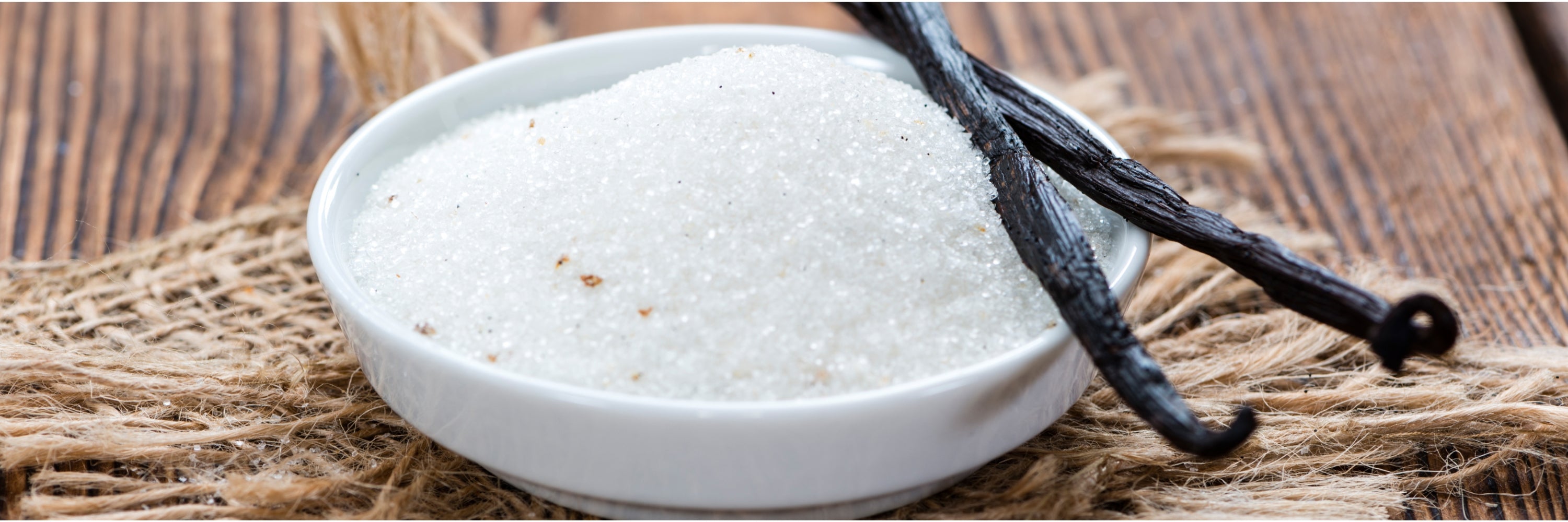 A small white bowl filled with granulated sugar sits on a burlap placemat, with two vanilla pods resting on the edge of the bowl.